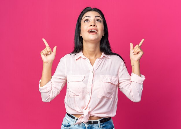 Junge schöne Frau in Freizeitkleidung, die glücklich und aufgeregt aufschaut und mit den Zeigefingern nach oben über rosa Wand zeigt