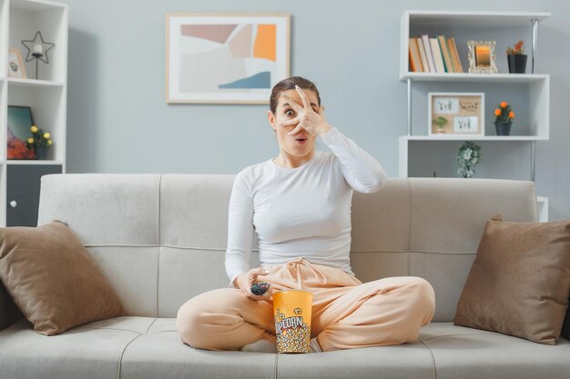 Junge schöne frau in freizeitkleidung, die zu hause auf einer couch sitzt, mit einem eimer popcorn, der einen gruseligen film in der hand hält, der verängstigt und besorgt aussieht und die augen mit einer handfläche bedeckt