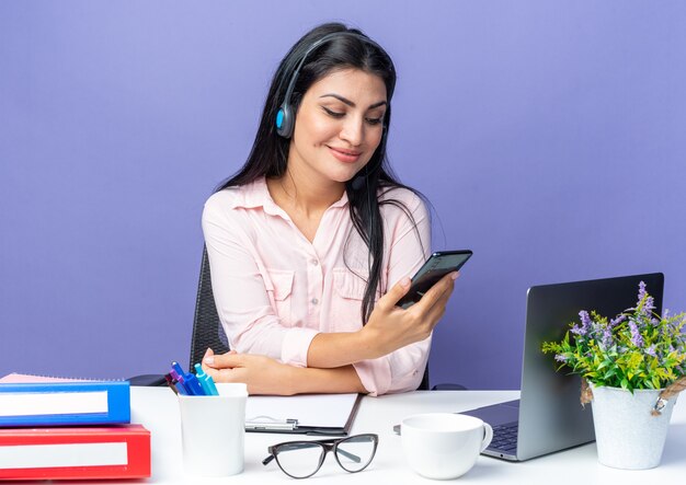 Junge schöne Frau in Freizeitkleidung mit Headset mit Smartphone und lächelnd am Tisch sitzend mit Laptop über blauer Wand im Büro arbeiten