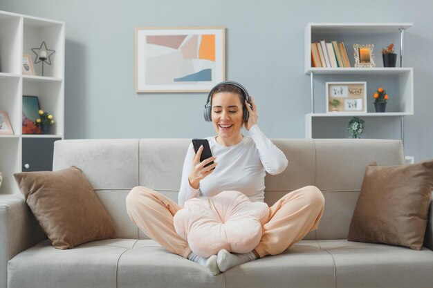 Junge schöne Frau in Freizeitkleidung mit Kopfhörern, die zu Hause auf einer Couch sitzt und ein Smartphone hält, das fröhlich glücklich und positiv lächelt und sich zu Hause entspannt