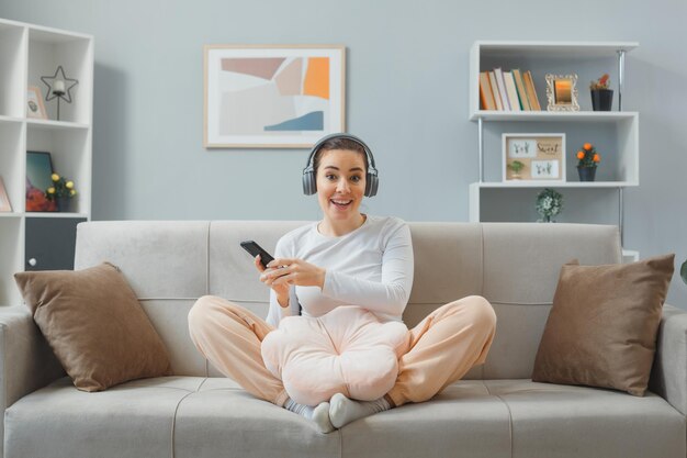 Junge schöne Frau in Freizeitkleidung mit Kopfhörern, die zu Hause auf einer Couch sitzt und ein Smartphone hält, das glücklich und positiv lächelnd in die Kamera blickt und sich entspannt zu Hause entspannt