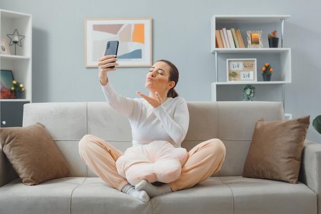 Junge schöne frau in freizeitkleidung sitzt auf einer couch zu hause und hält ein smartphone mit einem videoanruf, der glücklich und positiv einen luftkuss sendet, während sie sich zu hause entspannt