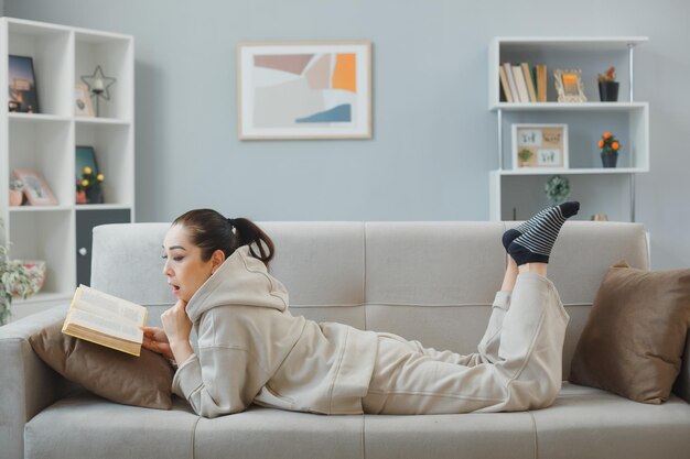 Junge schöne Frau in Hauskleidung, die zu Hause auf einer Couch liegt und erstaunt und überrascht aussieht, während sie ein Buch liest