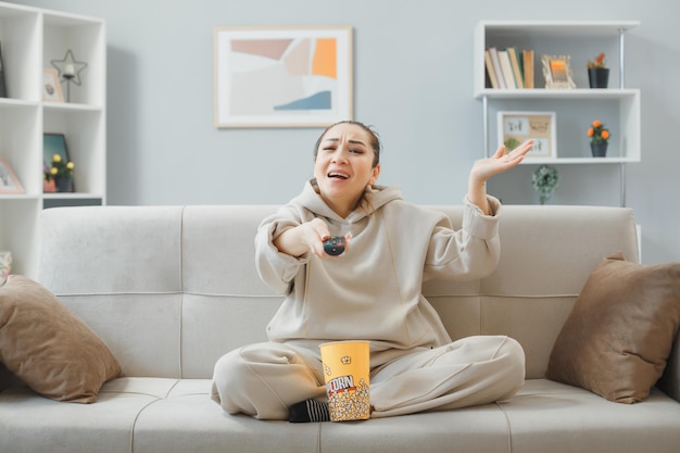 Junge schöne Frau in Hauskleidung, die zu Hause auf einer Couch sitzt, mit einem Eimer Popcorn, der die Fernbedienung beim Fernsehen hält und verwirrt ist, als sie den Arm vor Unmut hebt