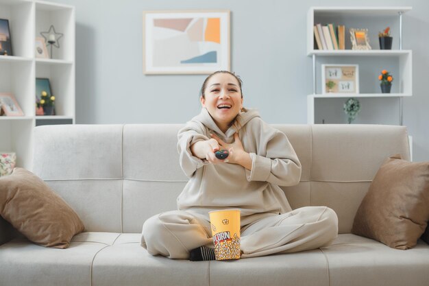 Junge schöne Frau in Hauskleidung, die zu Hause auf einer Couch sitzt, mit einem Eimer Popcorn, der die Fernbedienung hält und fernsieht, glücklich und fröhlich vor Lachen