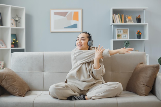 Junge schöne Frau in Hauskleidung, die zu Hause auf einer Couch sitzt, mit Kopfhörern, glücklich und positiv, sich entspannend, sich dehnend und Übungen machend