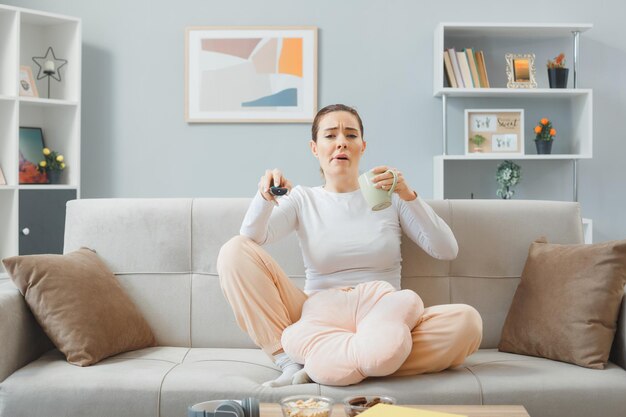 junge schöne frau in lässiger kleidung, die zu hause auf einer couch sitzt und eine fernbedienung und eine tasse tee hält, die verwirrt und unzufrieden aussieht, während sie fernsieht und das wochenende zu hause verbringt