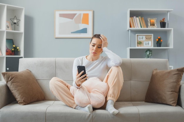 Junge schöne Frau in lässiger Kleidung, die zu Hause auf einer Couch sitzt und eine Smartphone-Lesenachricht hält, die müde und gelangweilt mit der Hand auf dem Kopf aussieht