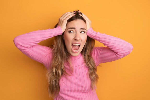 Junge schöne Frau in rosa Top, die frustriert schreit, wild wird und sich die Haare über Orange zieht