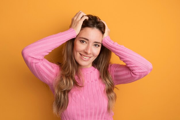 Junge schöne Frau in rosa Top, die glücklich und positiv in die Kamera schaut, mit den Händen auf dem Kopf, die über orangefarbenem Hintergrund stehen