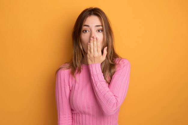 Junge schöne Frau in rosa Top, die schockiert in die Kamera schaut und den Mund mit der Hand bedeckt, die über orangefarbenem Hintergrund steht