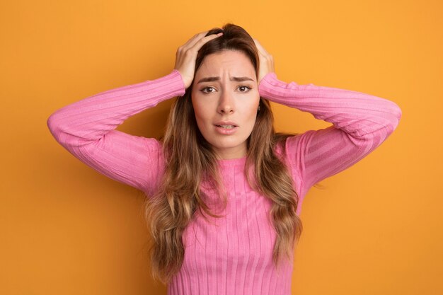 Junge schöne Frau in rosa Top, die verwirrt und sehr ängstlich in die Kamera schaut, mit den Händen auf dem Kopf, die über orangefarbenem Hintergrund stehen