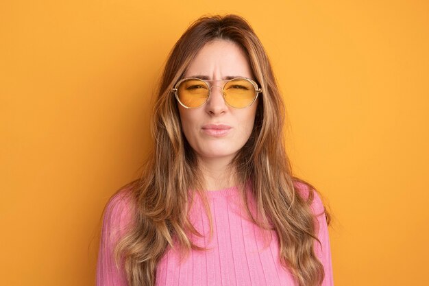 Junge schöne Frau in rosa Top mit Brille, die unzufrieden in die Kamera schaut, stirnrunzelnd über Orange stehend