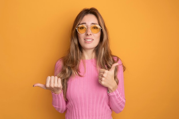 Kostenloses Foto junge schöne frau in rosa top mit brille, die verwirrt in die kamera schaut und daumen nach oben zeigt, die auf die seite zeigen, wobei der daumen über orangefarbenem hintergrund steht