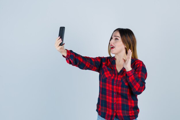 Junge schöne Frau macht Selfie, winkt mit der Hand zur Begrüßung im Freizeithemd und sieht glücklich aus. Vorderansicht.