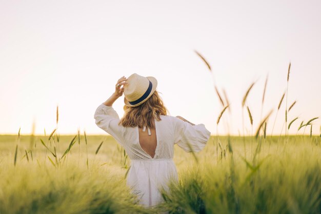 junge schöne frau mit blonden langen haaren in einem weißen kleid in einem strohhut sammelt blumen auf einem weizenfeld. Fliegende Haare in der Sonne, Sommer. Zeit für Träumer, goldener Sonnenuntergang.