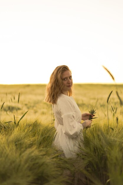 junge schöne frau mit blonden langen haaren in einem weißen kleid in einem strohhut sammelt blumen auf einem weizenfeld. Fliegende Haare in der Sonne, Sommer. Zeit für Träumer, goldener Sonnenuntergang.