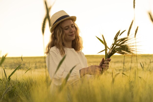 junge schöne frau mit blonden langen haaren in einem weißen kleid in einem strohhut sammelt blumen auf einem weizenfeld. Fliegende Haare in der Sonne, Sommer. Zeit für Träumer, goldener Sonnenuntergang.