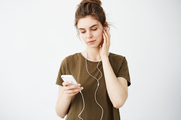 Junge schöne Frau mit Brötchen lächelnd, das Telefon betrachtet, das Musik in den Kopfhörern über weißem Hintergrund hört.