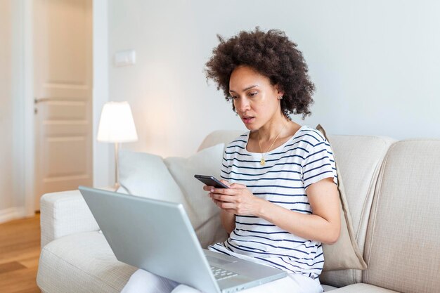 Junge schöne Frau mit dem lockigen Haar, das auf Laptop-Computer beim Sitzen auf dem Sofa zu Hause arbeitet