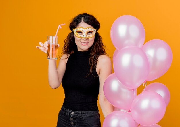 Junge schöne Frau mit dem lockigen Haar, das Bündel Luftballons in der Parteimaske glücklich und fröhlich trinkt Cocktails Geburtstagsfeierkonzept steht über orange Wand hält