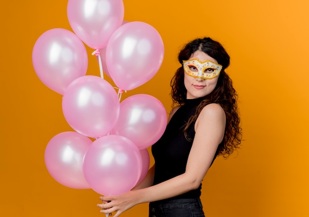 Junge schöne Frau mit dem lockigen Haar, das Bündel Luftballons in der Partymaske glücklich und fröhlich Geburtstagsfeierkonzept über Orange hält