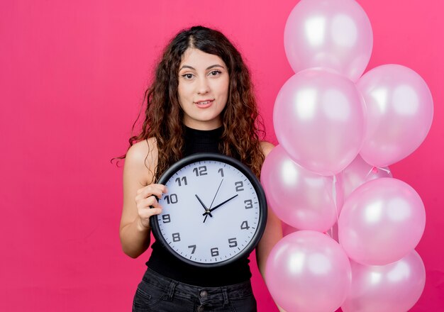 Kostenloses Foto junge schöne frau mit dem lockigen haar, das bündel luftballonwanduhr lächelt, die fröhlich über rosa wand steht