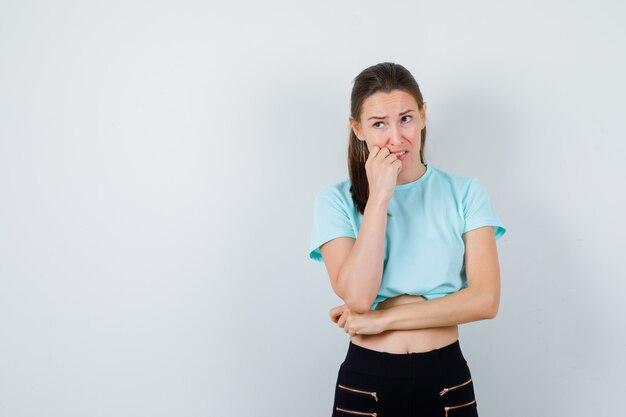 Junge schöne Frau mit der Hand im Gesicht, wegschauen im T-Shirt, in der Hose und nachdenklich. Vorderansicht.