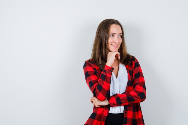 Junge schöne Frau mit der Hand unter dem Kinn, die im lässigen Outfit wegschaut und glückselig aussieht, Vorderansicht.