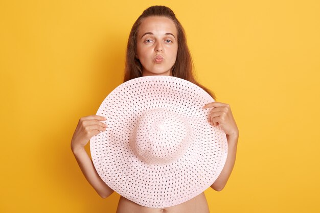 Junge schöne Frau mit dunklem Haar ohne Kleidung, versteckt sich hinter großem Strohhut, der über gelber Wand isoliert wird, und hält Lippen gerundet, wie Kussgeste zeigend.