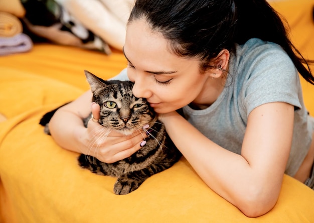 Junge schöne Frau mit Katze zu Hause, die auf dem orangefarbenen Sofa sitzt und ihr niedliches Haustier umarmt