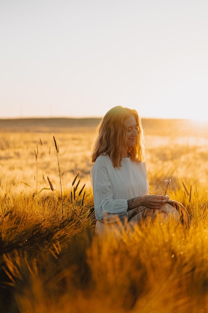 junge schöne Frau mit langen blonden Haaren in einem weißen Kleid auf einem Weizenfeld am frühen Morgen bei Sonnenaufgang. Der Sommer ist die Zeit für Träumer, fliegende Haare, eine Frau, die in den Strahlen über das Feld rennt