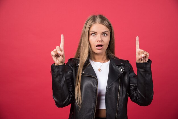 Junge schöne Frau mit Lederjacke, die über roter Wand steht und mit den Fingern nach oben zeigt