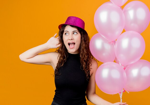 Junge schöne Frau mit lockigem Haar in einem Feiertagshut, der Bündel Luftballons glücklich und aufgeregt lächelt fröhlich Geburtstagsfeierkonzept, das über orange Wand steht