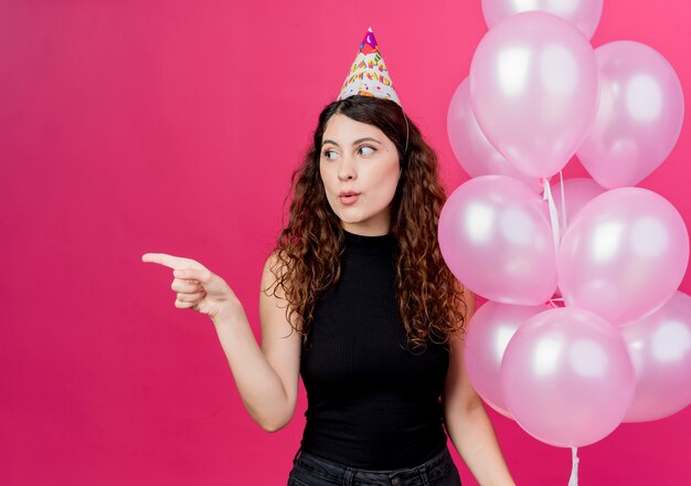 Junge schöne Frau mit lockigem Haar in einer Feiertagskappe, die Bündel Luftballons hält, die mit Finger auf das Seitengeburtstagsfeierkonzept zeigen, das über rosa Wand steht