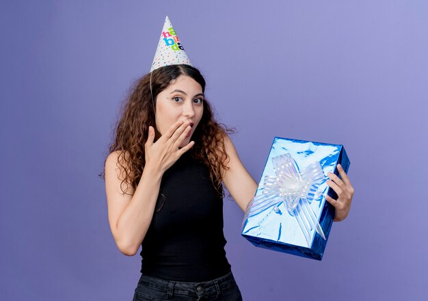 Junge schöne Frau mit lockigem Haar in einer Feiertagskappe, die Geburtstagsgeschenkbox hält, die überraschtes Geburtstagsfeierkonzept steht, das über blauer Wand steht