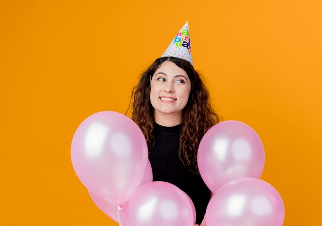 Junge schöne Frau mit lockigem Haar in einer Feiertagskappe, die glückliche und aufgeregte Geburtstagsfeierkonzept der Luftballons hält, die über orange Wand stehen