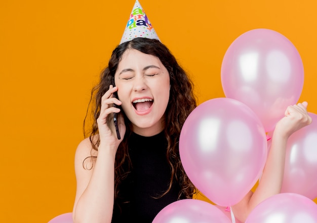 Junge schöne Frau mit lockigem Haar in einer Feiertagskappe, die Luftballons hält, die auf dem glücklichen und aufgeregten Geburtstagsfeierkonzept des Mobiltelefons über orange Wand stehen