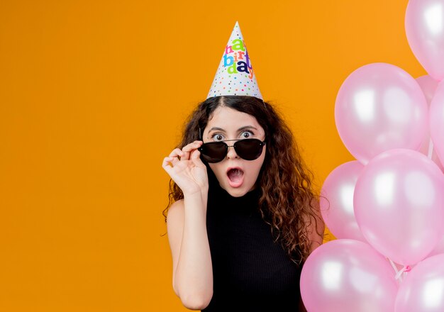 Junge schöne Frau mit lockigem Haar in einer Feiertagskappe, die Luftballons hält, die Gläser abnehmen, die überraschtes Geburtstagsfeierkonzept stehen, das über orange Wand steht