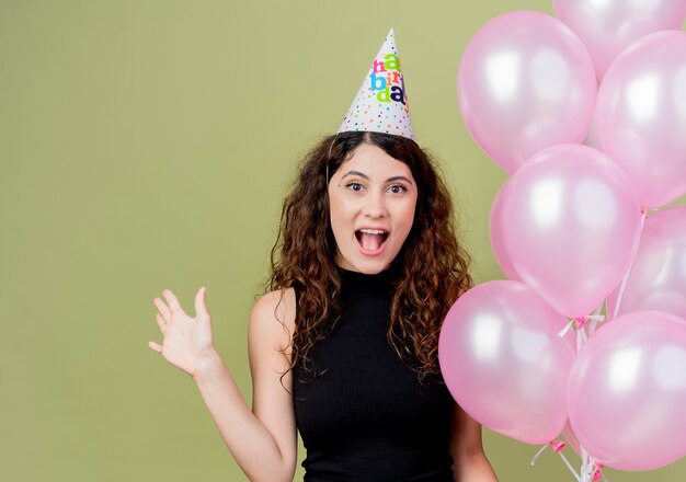 Kostenloses Foto junge schöne frau mit lockigem haar in einer feiertagskappe, die luftballons hält, die glückliches und aufgeregtes lächelndes fröhliches geburtstagsfeierkonzept der hand anheben, das über lichtwand steht