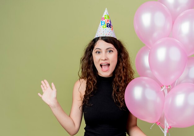 Junge schöne Frau mit lockigem Haar in einer Feiertagskappe, die Luftballons hält, die glückliches und aufgeregtes lächelndes fröhliches Geburtstagsfeierkonzept der Hand anheben, das über Lichtwand steht