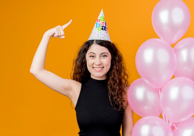 Junge schöne Frau mit lockigem Haar in einer Feiertagskappe, die Luftballons hält, die glückliches und positiv lächelndes Geburtstagsfeierkonzept des Zeigefingers zeigen, das über orange Wand steht