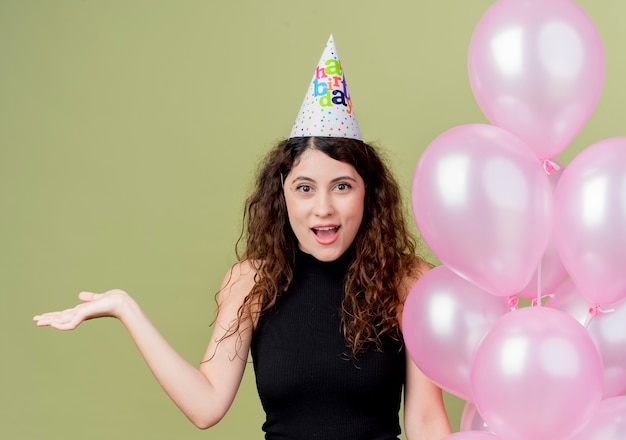 Junge schöne Frau mit lockigem Haar in einer Feiertagskappe, die Luftballons hält, die überrascht und glücklich feiern Geburtstagsfeier über Lichtwand stehen