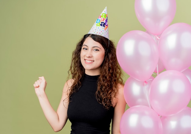 Junge schöne Frau mit lockigem Haar in einer Feiertagskappe, die Luftballons hält geballte Faust glückliches und aufgeregtes Geburtstagsfeierkonzept, das über Lichtwand steht