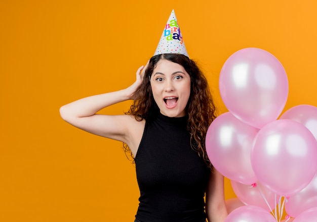 Junge schöne Frau mit lockigem Haar in einer Feiertagskappe, die Luftballons überrascht und alles Gute zum Geburtstagfeierkonzept hält, das über orange Wand steht