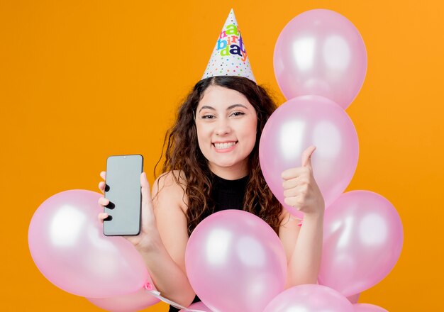 Junge schöne Frau mit lockigem Haar in einer Feiertagskappe, die Luftballons und Smartphone hält, die Daumen oben glückliches und aufgeregtes Geburtstagsfeierkonzept über Orange zeigen