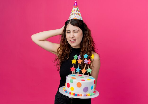 Junge schöne Frau mit lockigem Haar in einer Feiertagsmütze, die Geburtstagstorte hält, die mit Hand auf ihrem Kopfgeburtstagsfeierkonzept verwirrt steht, das über rosa Wand steht