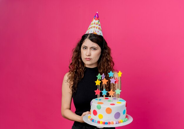Junge schöne Frau mit lockigem Haar in einer Feiertagsmütze, die Geburtstagstorte hält, missfiel mit Stirnrunzeln Gesicht Geburtstagsfeier Konzept über rosa Wand