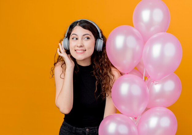 Junge schöne Frau mit lockigem Haar mit Kopfhörern, die Musik hören, die Bündel Luftballons glückliches und fröhliches Geburtstagsfeierkonzept hält, das über orange Wand steht