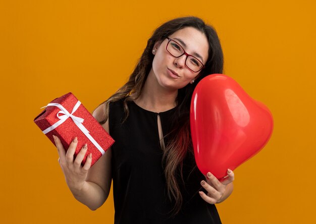 Junge schöne Frau mit rotem Ballon in Herzform und Geschenk glücklich und fröhlich lächelnd zum Valentinstag